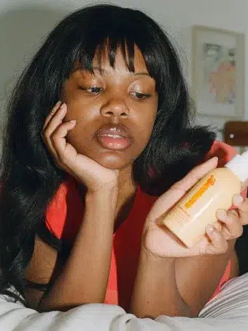 Person in a red shirt lies on a bed, looking at a bottle of skincare product.