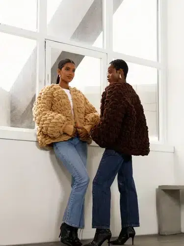 Two individuals in textured jackets, one beige and one dark brown, stand by a large window. They wear jeans and black heeled boots, facing each other.