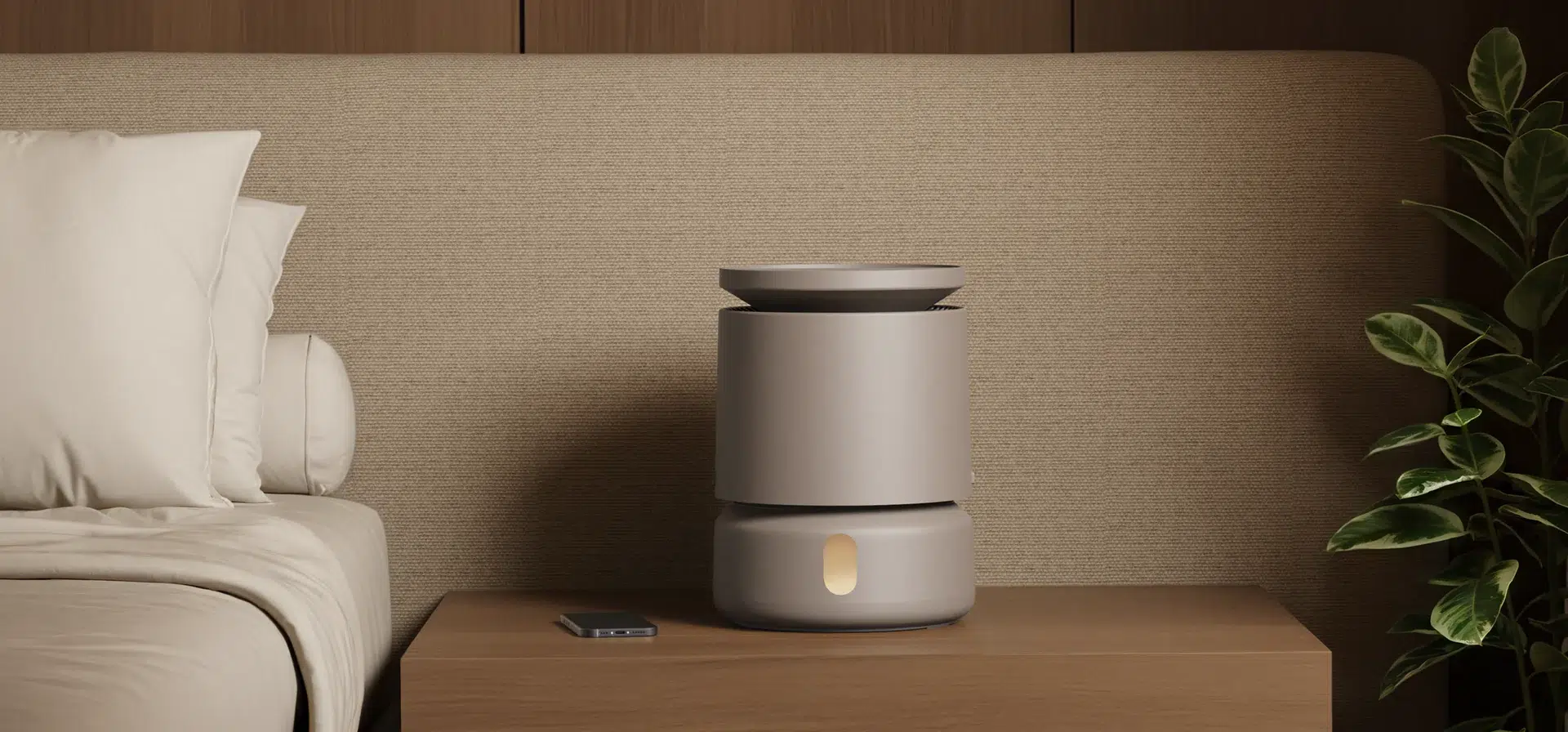 A cylindrical, modern air purifier sits on a wooden nightstand next to a bed, with a plant and a remote control also on the table.