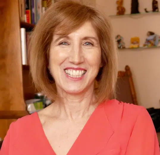 A woman with shoulder-length brown hair smiles while wearing a coral top. Shelves and a wooden bookcase are in the background.