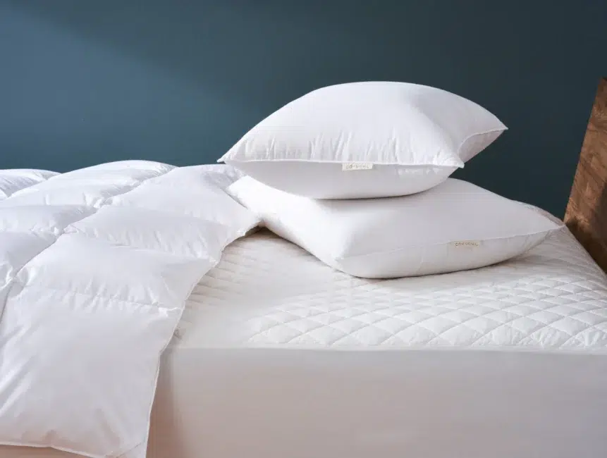 Two white pillows stacked on a bed with a quilted mattress cover and a folded white comforter, against a dark blue wall.