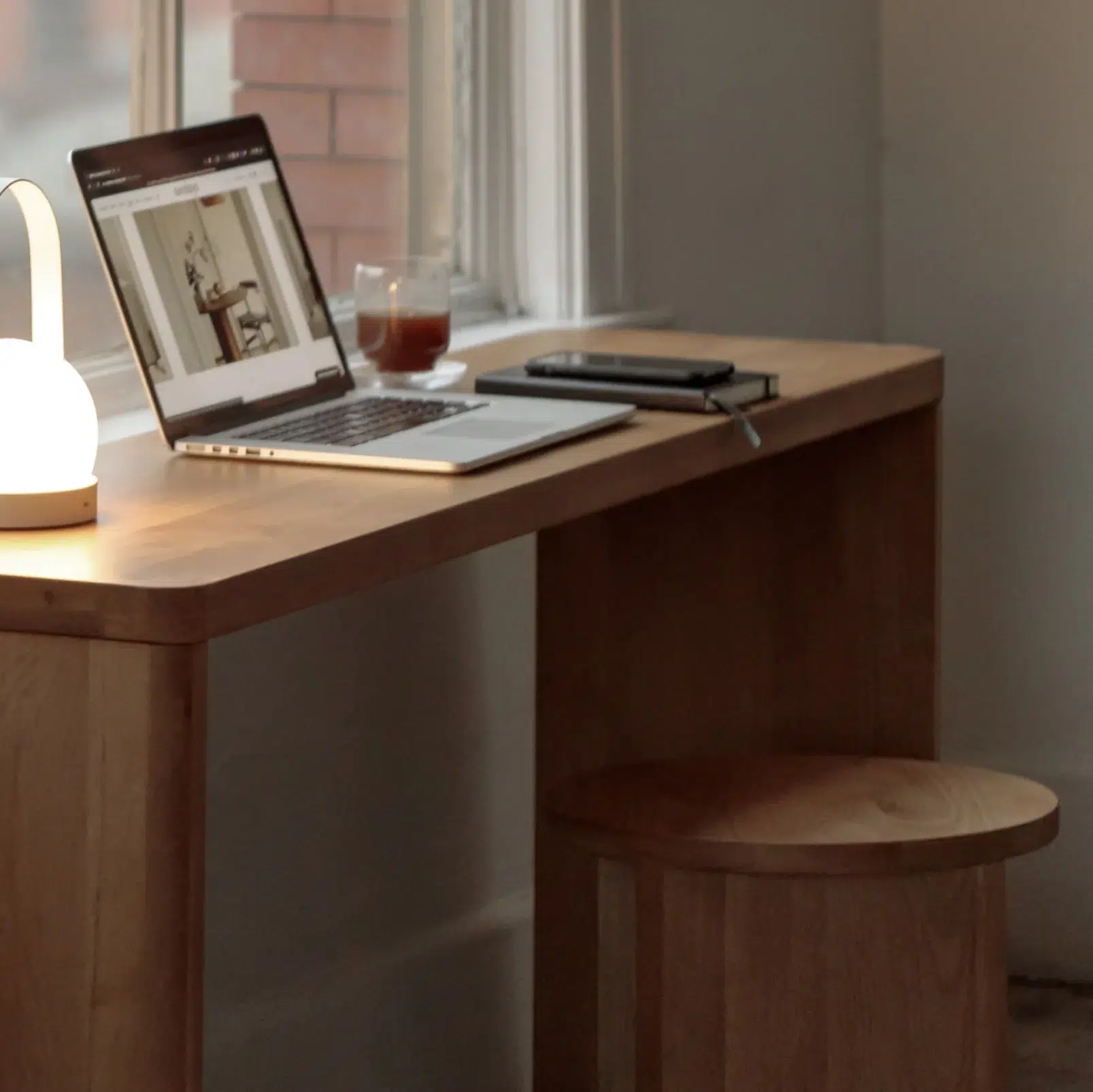 A minimalist wooden desk by a window with a lit lamp, an open laptop, a glass of drink, a phone, and a closed notebook on its surface, with a matching wooden stool.