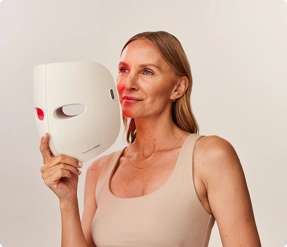 A woman holds a white LED light therapy mask to her face, wearing a beige tank top against a neutral background.