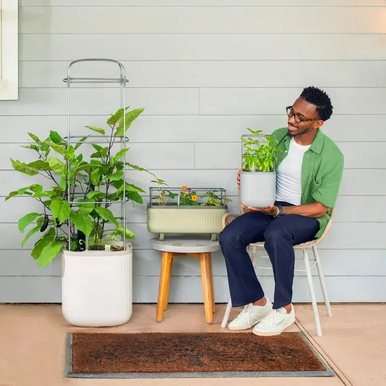 A person sits on a chair holding a small potted plant, with two larger planters and a small planter arranged on a stool beside them against a paneled wall.