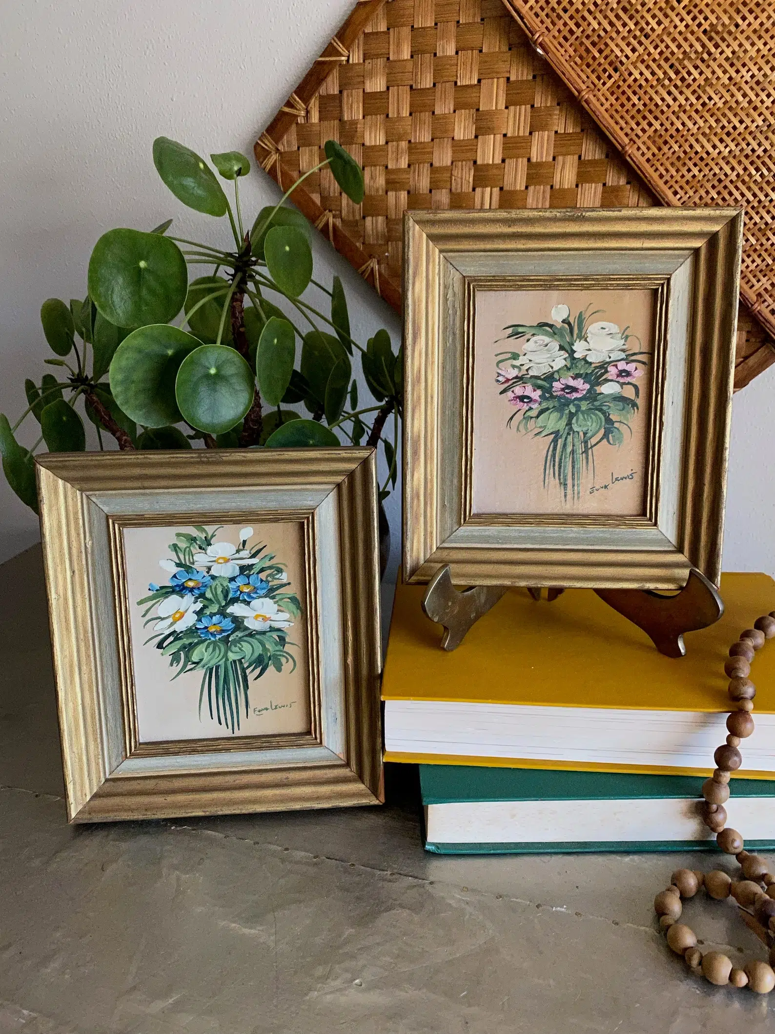 Two small framed floral paintings on a table, accompanied by stacked books, a beaded necklace, and a potted plant.
